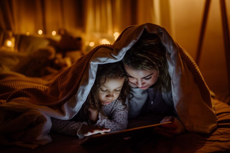 Happy mother with her little daughter lying under blanket and watching movie on tablet at home