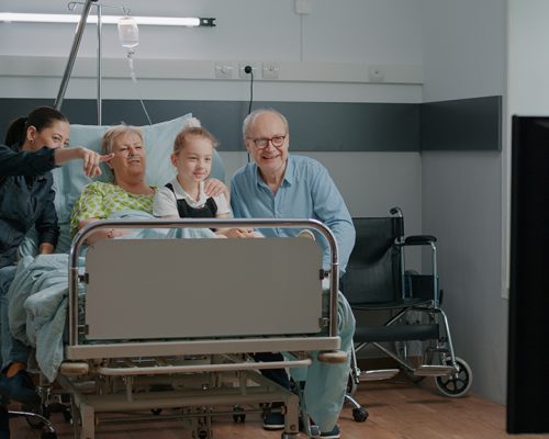 Family watching television and visiting senior patient in hospital ward Family watching television and visiting senior patient in hospital ward. Elder woman enjoying visit from child and daughter, being hospitalized to cure disease. People having fun with tv.