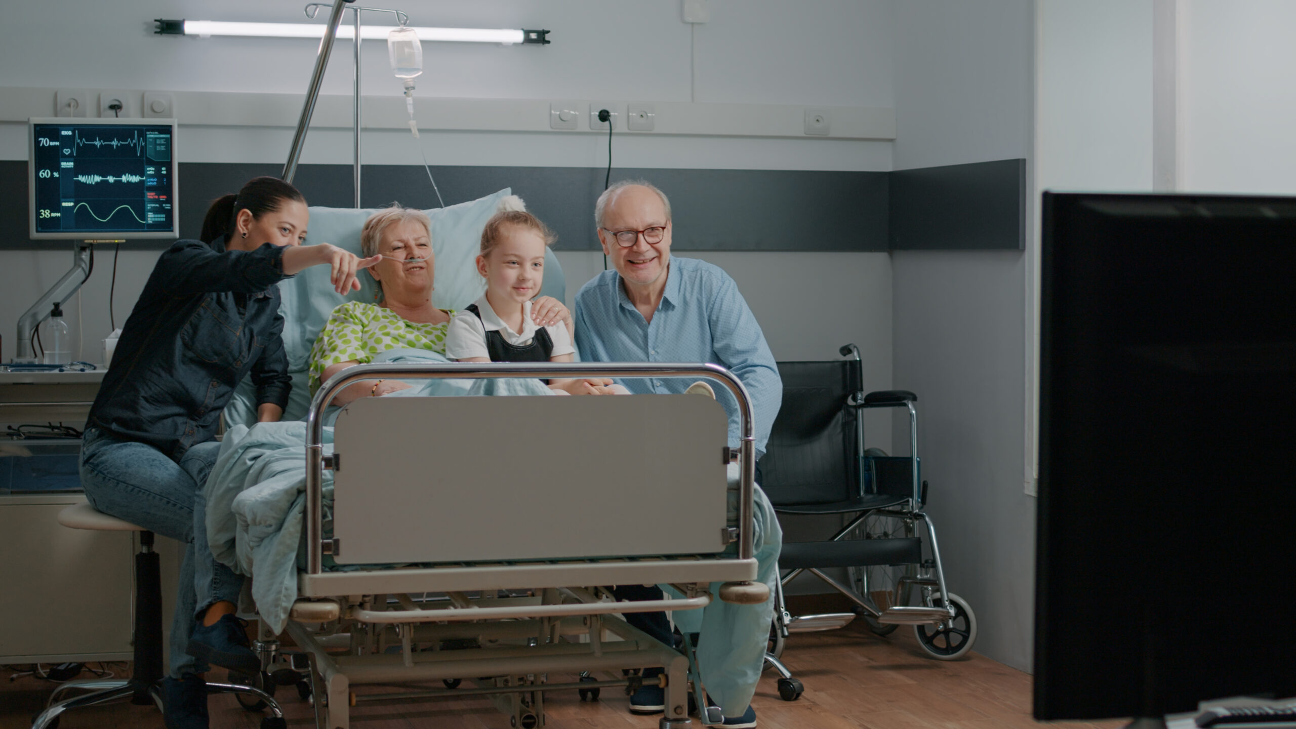 Family watching television and visiting senior patient in hospital ward. Elder woman enjoying visit from child and daughter, being hospitalized to cure disease. People having fun with tv.